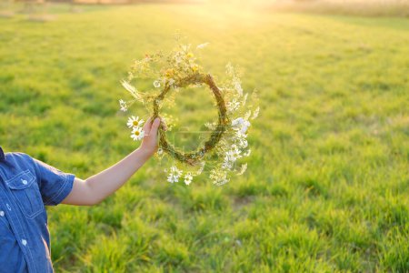 happy boy holding wreath in hand, floral crown on green sunlit meadow, seen from behind, beauty nature and arrival summer, Midsummer celebration, wiccan Litha sabbat, pagan holiday