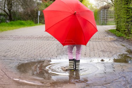 Foto de Niña traviesa de 5 años de edad, con botas de goma con paraguas rojo se para en el charco de lluvia, la captura de alegrías puras y simples infancia y magia día lluvioso - Imagen libre de derechos