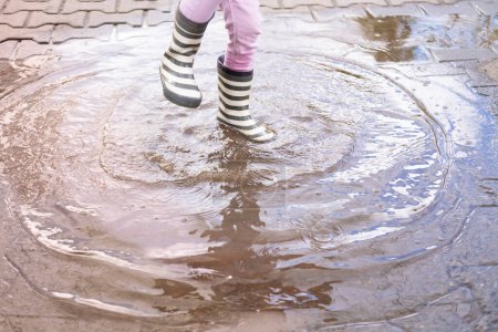 Foto de Primer plano niña de 5 años salta alegremente en charco con botas de goma, pies de los niños en salpicaduras de agua, saltos felices, placeres de la infancia - Imagen libre de derechos