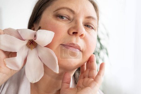 Foto de Mujer envejecida con flor de magnolia se mira en el espejo, notando arrugas, cambios en el contorno facial y párpados caídos, procedimientos cosméticos anti-envejecimiento, proceso natural envejeciendo - Imagen libre de derechos