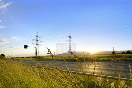 beautiful dramatic landscape, car road visible behind grass, power towers, field in evening in rays sunset, concept beauty of nature, modern energy, technology, seasonal change