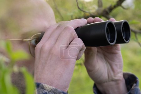 young man, hidden in lush greenery spies clad peers through binoculars, private investigator conducts surveillance, thrill-seeker tracks down target