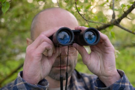 young man, hidden in lush greenery spies clad peers through binoculars, covert operation, thrill-seeker tracks down target