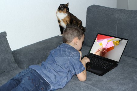 Téléchargez les photos : Enfant intelligent 8-10 ans dans les écouteurs se trouve sur le canapé devant l'ordinateur portable, regarde le blog des enfants sur l'origami, artisanat artisanal, concept d'enseignement à distance, apprentissage, auto-éducation - en image libre de droit