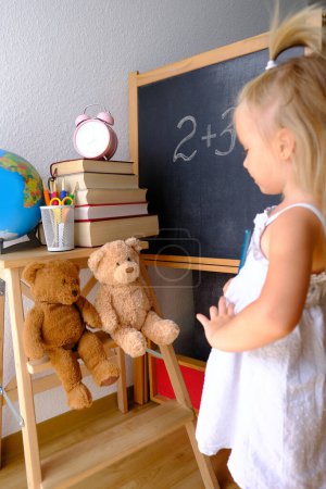 child, blonde girl plays school, small teacher teaches lesson, student home office, alarm clock, books, teddy bears, chalk board, globe, concept of education, back to school, knowledge day