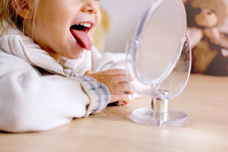 Blondes Mädchen von 3 Jahren vor dem Spiegel beschäftigt mit Logopäde, Zungengymnastik, Defekt, Sprachstörung mit häufigen Wiederholungen Laute, Silben, Krämpfe Muskelapparat