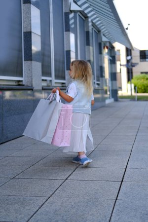 happy child of 3-4 years old go out of store, girl in light fashionable clothes, craft paper bags full of purchases in both hands, happy shopping on Black Friday, young shopaholic, little fashion-girl