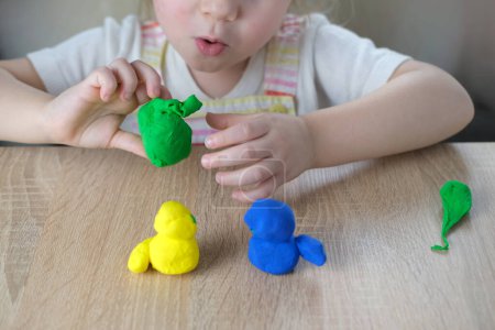 close-up plasticine toys in hands of toddler, small child, blonde girl 2 years old sculpts figures of characters from colored dough, concept of fine motor skills, tactile sensations, creativity