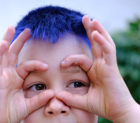 child on natural green background, boy 8-9 years with blue hair smiles cheerfully, upper part of face, concept of cheerful childhood, self-identification, desire to stand out from crowd of peers