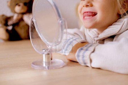 blonde girl of 4 years old girl in front of mirror engaged with speech therapist, gymnastics for tongue, defect, speech disorder with frequent repetition sounds, syllables, spasms muscles apparatus