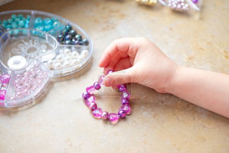 Preschool girl engaged in arts and crafts activity, DIY craft project, making personalized bracelet with colorful beads, fine motor skills, promoting creativity and self-expression