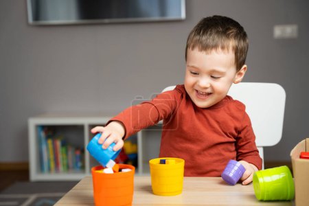 A happy smiling cute little toddler boy of two years old sits at a children's table and plays with multi-colored pyramids in his room. Educational toys for children. Soft selective focus