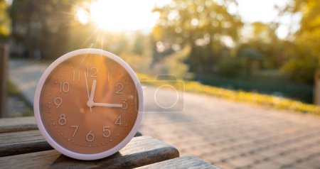 Foto de El reloj está parado en un banco en el parque en un día soleado de otoño. Cambiar el concepto de tiempo. Planificar su día y administrar el tiempo. Otoño temporada de otoño. DST. Enfoque selectivo - Imagen libre de derechos