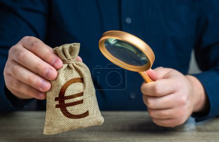 Téléchargez les photos : Un homme examine un sac en euros avec une loupe. Contrôle de l'origine du capital. Lutte contre le blanchiment d'argent et l'évasion fiscale. Modalités de l'étude en dépôt ou en prêt. Trouver des investissements - en image libre de droit