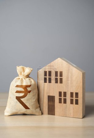Wooden house figure and indian rupee money bag. Taxes. Property value appraisal. Make a deal. Property Insurance. Housing prices. Buying and selling real estate.