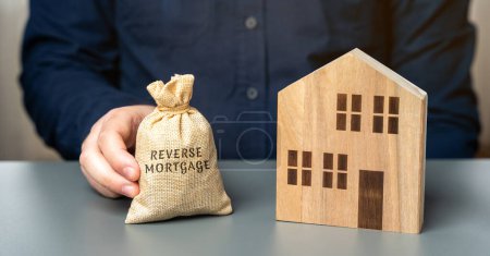 Photo for Reverse mortgage concept. Allowing homeowners to borrow money by using their home as collateral for the loan. Money bag in the hands of a businessman and a wooden house - Royalty Free Image