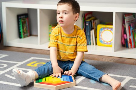 Toddler boy with a bandage or cast on his leg plays with wooden blocks. Fracture of a foot and finger in children. Human healthcare and medicine concept