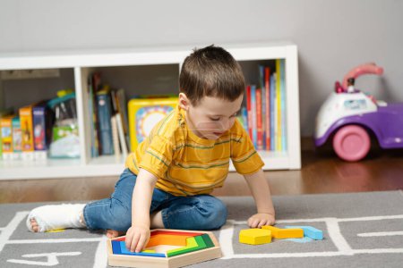 El niño pequeño con un vendaje o un yeso en la pierna juega con bloques de madera. La fractura del pie y el dedo en los niños. Salud humana y concepto de medicina