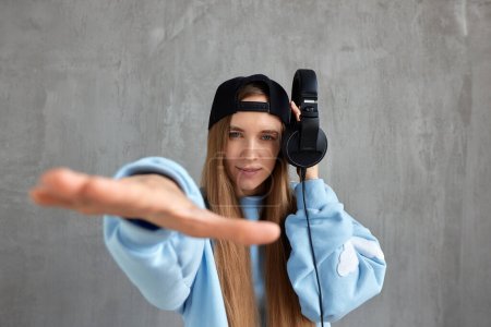 Photo for A young pretty long-haired DJ girl in a blue sweater and a black baseball cap holds headphones in her hand and imitates the movements of a hip-hop musician. Music, fun and beauty - Royalty Free Image