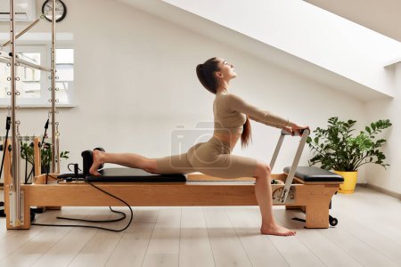 Téléchargez les photos : Une jeune fille fait du Pilates sur un lit réformateur dans un studio lumineux. Une belle brunette élancée en body beige fait des exercices pour renforcer son corps et étirer ses jambes - en image libre de droit