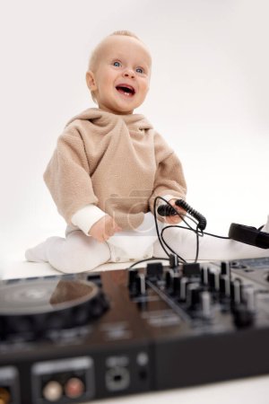 Foto de Un niño sonriente lindo en una sudadera con capucha beige está sentado en el suelo con auriculares dj y un tablero de mezcla dj. Música y diversión. Aislado sobre fondo blanco - Imagen libre de derechos