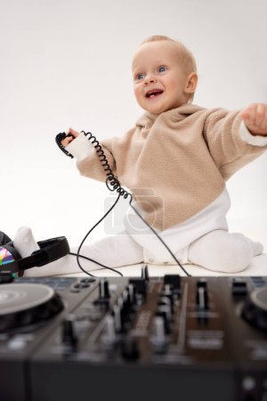 Photo for A cute smiling little child in a beige hoodie is sitting on the floor with dj headphones and a dj mixing board. Music and fun. Isolated on white background - Royalty Free Image