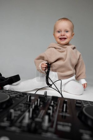 Photo for A cute smiling little child in a beige hoodie is sitting on the floor with dj headphones and a dj mixing board. Music and fun. Isolated on grey background - Royalty Free Image