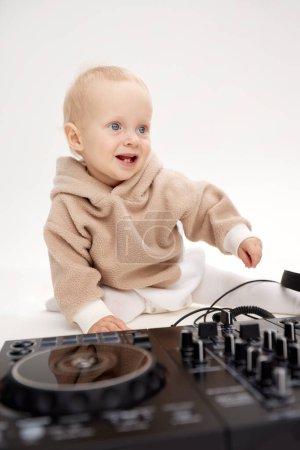 Photo for A cute smiling blonde baby in a beige hoodie sits on the floor and plays with dj headphones and a dj mixing board. Music and fun. Isolated on a white background - Royalty Free Image