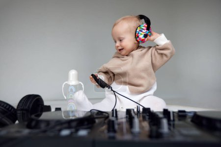 Photo for A cute smiling blonde baby in a beige hoodie sits on the floor and plays with dj headphones and a dj mixing board. Music and fun. Isolated on a gray background - Royalty Free Image