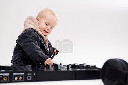 Photo for A cute kid in a beige hoodie and a funny black motorcycle jacket sits on the floor with dj headphones and a dj mixing console. Music and fun. Isolated on a white background - Royalty Free Image