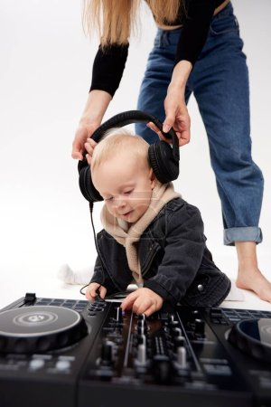 Photo for A cute kid in a beige hoodie and a funny black motorcycle jacket sits on the floor with a dj mixing console. A barefoot mom in blue jeans puts DJ headphones on his head. - Royalty Free Image