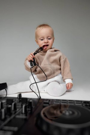 Foto de Un lindo bebé en una sudadera con capucha beige se sienta en el suelo y juega con auriculares DJ y una consola de mezcla de DJ. Música y diversión. Aislado sobre un fondo gris - Imagen libre de derechos