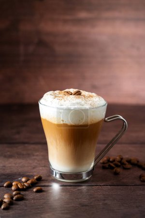 Glass cup of layered coffee drink with milk foam, coffee beans, wooden background and copy space.