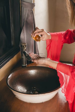 Photo for A woman in a red silk robe pouring hand gel from bottle into hand at night. - Royalty Free Image