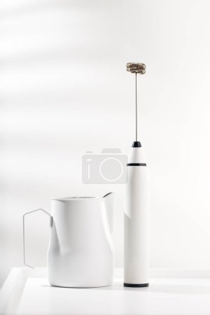 Milk frother and white pitcher for coffee on white minimalist table.