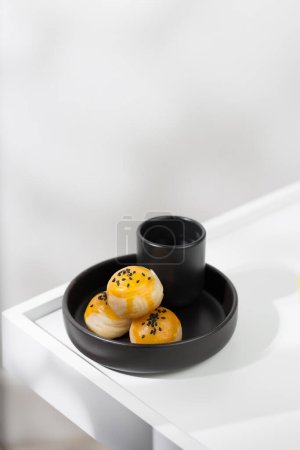 Téléchargez les photos : Pâtisserie chinoise saupoudrée de graines de sésame noir sur le dessus, et servie avec du thé chaud dans une tasse en céramique noire. - en image libre de droit