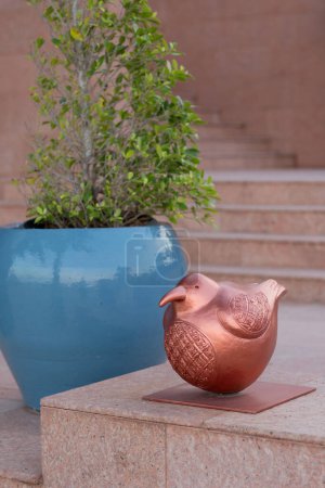 An elegant copper bird sculpture, intricately designed, rests on a stone surface. Adjacent, a large blue pot houses a lush green plant. The serene outdoor setting, featuring stone stairs, adds a tranquil ambiance to this still life composition.