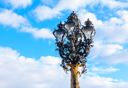 Téléchargez les photos : Lampadaire contre le ciel bleu avec des nuages. La base de la lampe présente des volutes et des courbes élaborées, caractéristiques de la période baroque - en image libre de droit