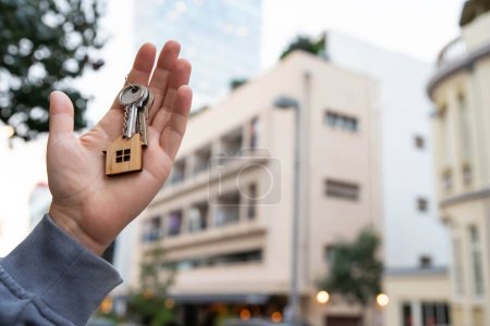 Buying a house, building repair and mortgage concept. Estimation real estate property with loan money and banking. Keys with toy home keychain charm in hand on city building background.