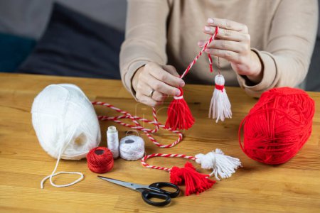 Photo for Woman making handmade traditional martisor, from red and white strings with tassel. Symbol of holiday 1 March, Martenitsa, Baba Marta, beginning of spring in Romania, Bulgaria, Moldova - Royalty Free Image