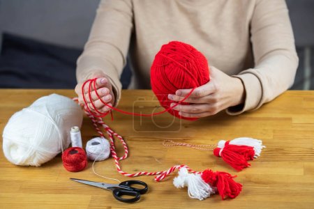 Photo for Woman making handmade traditional martisor, from red and white strings with tassel. Symbol of holiday 1 March, Martenitsa, Baba Marta, beginning of spring in Romania, Bulgaria, Moldova - Royalty Free Image