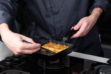 Foto de Chef hombre manos cocina Tamagoyaki o Tamago tradicional japonés Rolled Tortilla receta con palillos, hecho por rodar varias capas de huevo revuelto cocido en una sartén en una tortilla rectangular. - Imagen libre de derechos