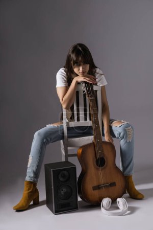 Photo for Young woman holdilng classic guitar sitting on a chair. Musician in studio near acoustic guitar and music column - Royalty Free Image