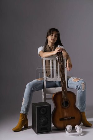 Photo for Young woman holdilng classic guitar sitting on a chair. Musician in studio near acoustic guitar - Royalty Free Image