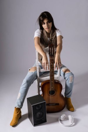 Photo for Young woman holdilng acoustic guitar sitting on a chair. Musician in studio with classic guitar - Royalty Free Image