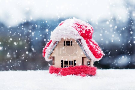 A wooden toy house against the backdrop of nature is covered with a hat and wrapped in a scarf in winter during snowfall. The concept of keeping warm