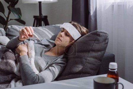 Woman under plaid feel sick unwell. Unhealthy young female measuring body temperature with electronic thermometer, suffer from flu lying on sofa at home, seasonal contagious grippe illness concept