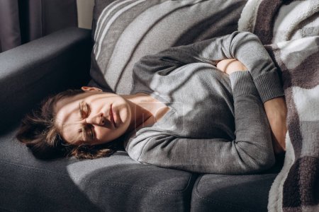 Depressive junge kaukasische Frau, die mit den Armen um sich herum auf dem Sofa liegt, unglückliche kaukasische Brünette, die an Schlaflosigkeit, psychischen Problemen, Konzeptimpotenz, Asthenie leidet