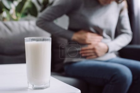 Photo for Lactose intolerance, food allergy concept. Selective focus of glass milk is on white table and unhealthy young female touch stomach suffers from severe ache sitting on couch in living room at home - Royalty Free Image