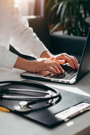 Female doctor in white coat with stethoscope using laptop, writing in medical journal, professional therapist practitioner sitting at table in hospital and typing at computer. Medicine concept